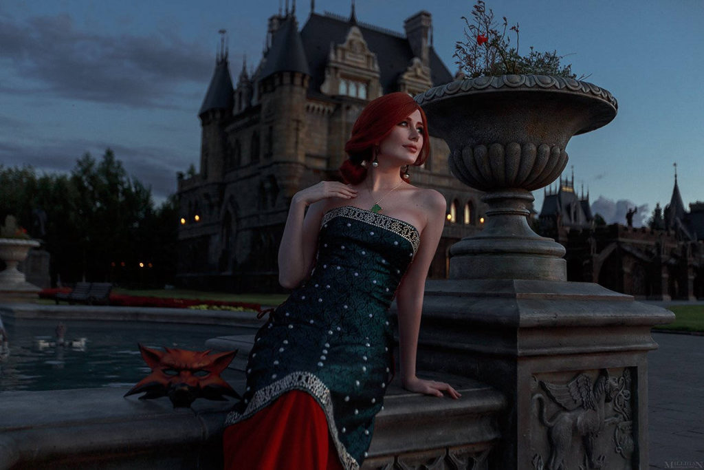 Wiedźmin 3 Dziki Gon cosplay - Triss - Vick_Torie jako Triss siedzi na murku fontanny i zalotnie patrzy się w dal, w tle widać zamek